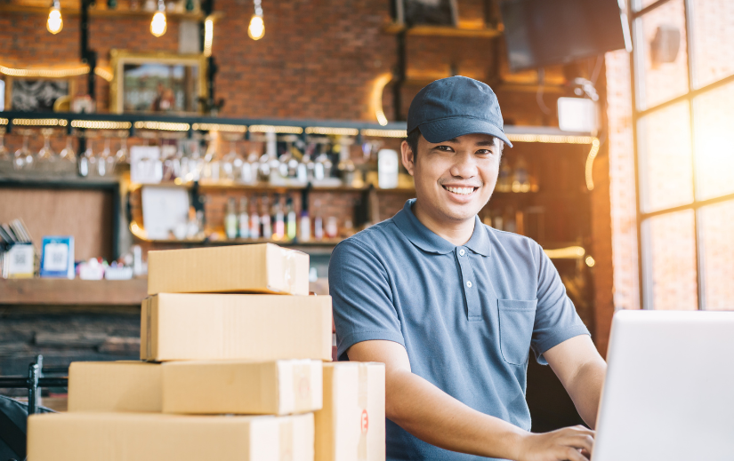 Smiling entrepreneur using a laptop with stacked packages, perfect for an Amazon beginner’s guide to starting online.